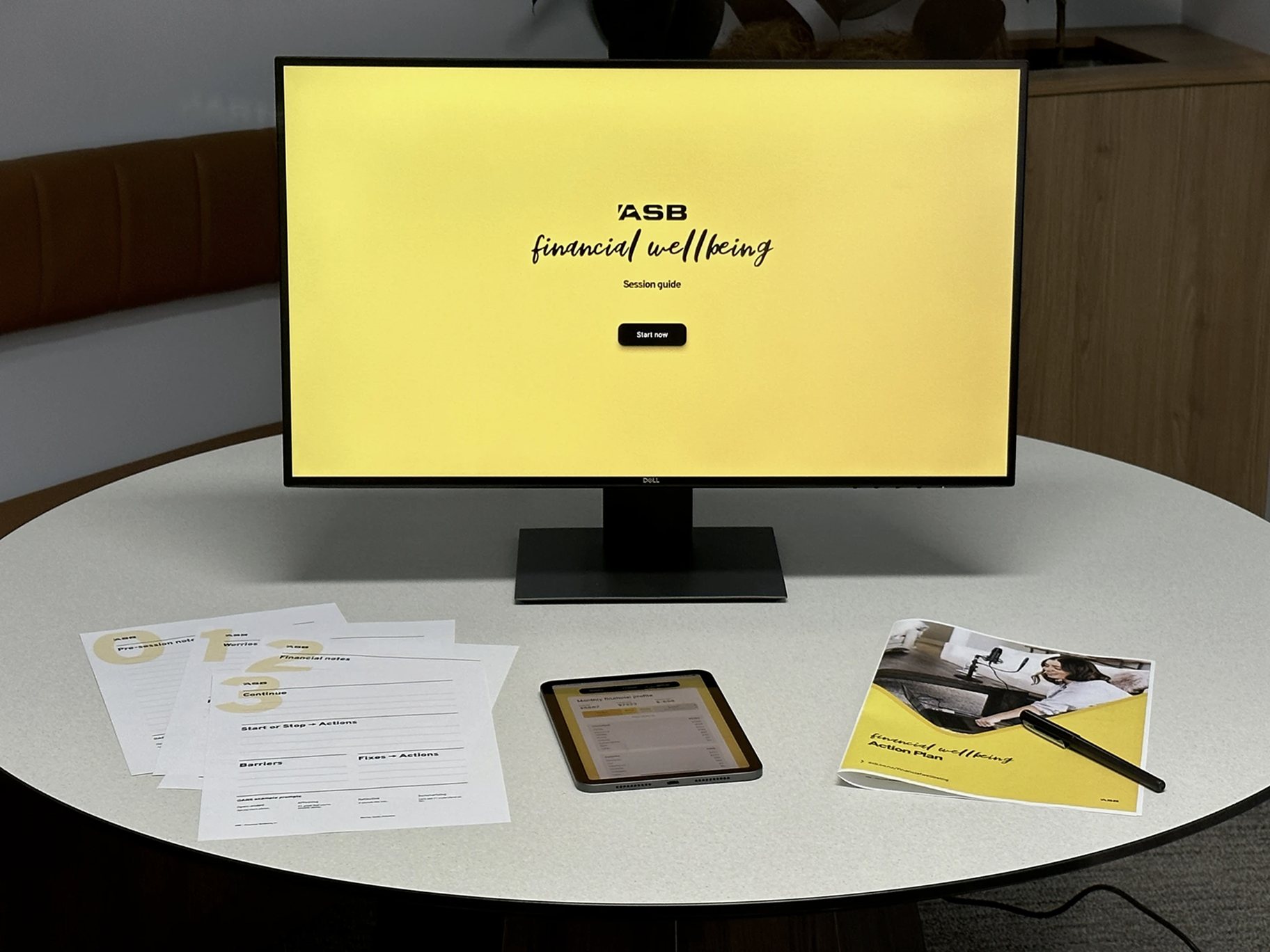 A table with a screen, an iPad and a printed booklet, and worksheets — all sporting Financial Wellbeing branding