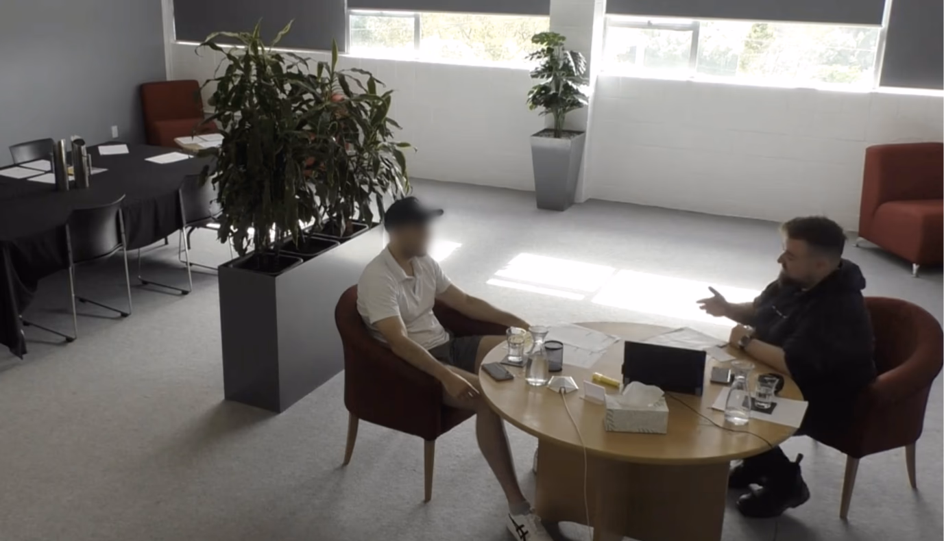 A circle table with 2 men sitting opposite, talking to one another.